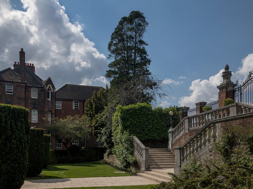 A landscaped garden featuring a paved walkway leading to stone steps bordered by an ornate balustrade and decorative columns, with lush green hedges and shrubs on either side. Tall trees, including a 